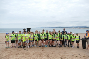 A large group of children in high-vis vests, smile for the camera. Alongside Sally, teacher Sam and a a parent.