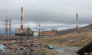 A picture of Norilsk Nickel plant in Siberia showing bare hillsides and large industrial buildings with red and white chimneys and large electrical pylons in Norilsk, Siberia