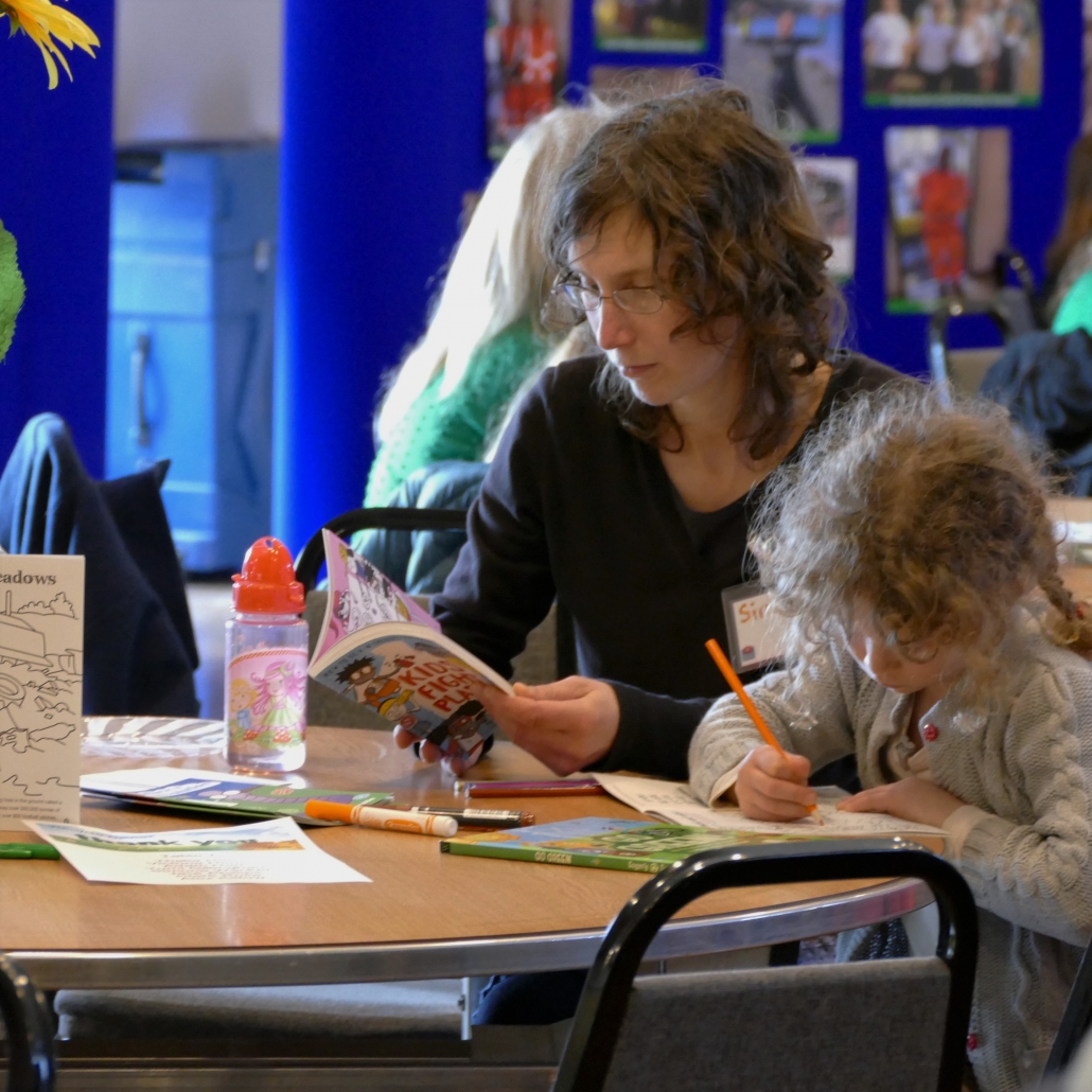 A young girl and a woman are sat at a table. The woman is reading a book while the young girl colours a page in a colouring book. There are colouring pens, books and leaflets strewn across the table.