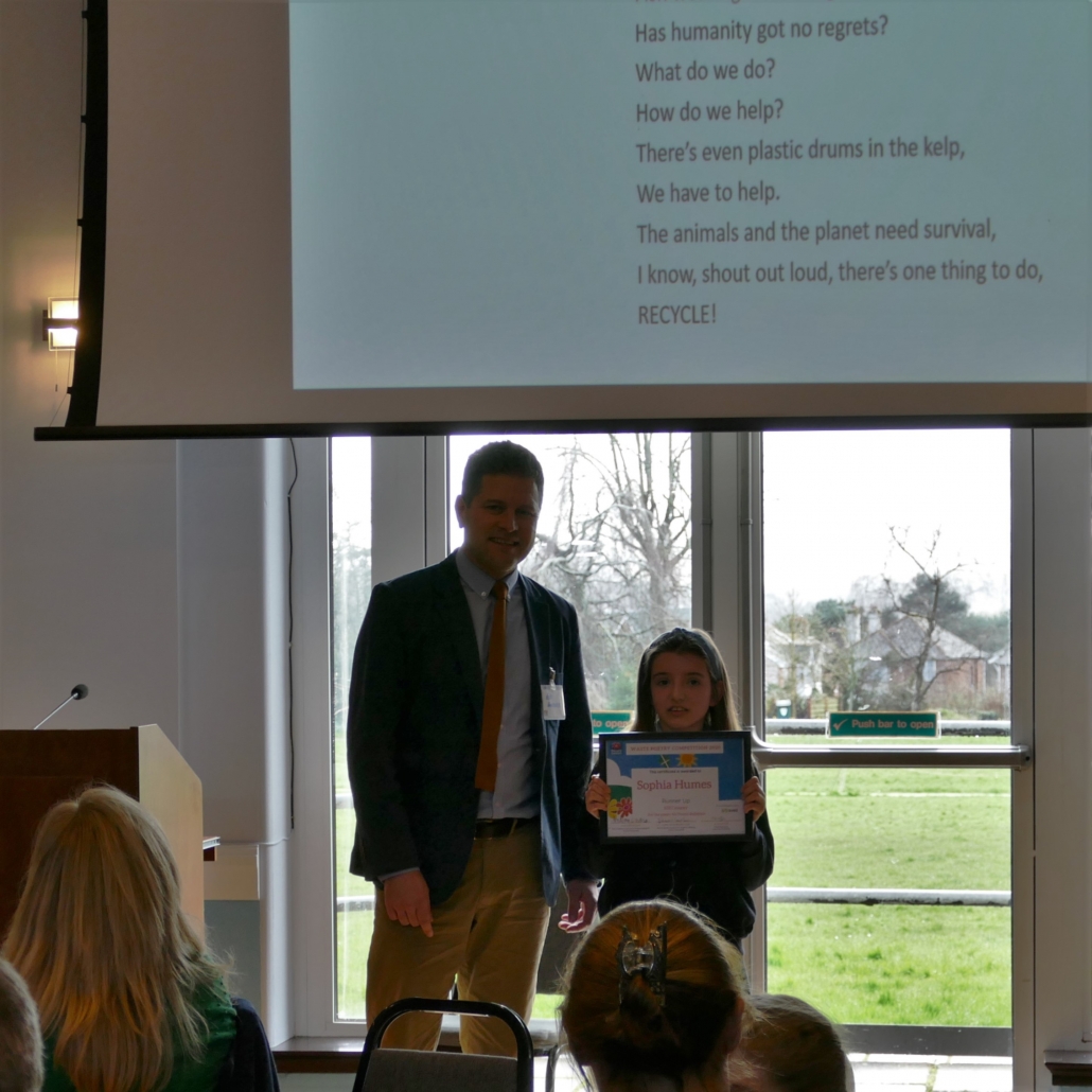 Rob Sanders standing alongside Sophia Humes, after presenting her with an award. Sophia's poem is displayed on a screen behind them.