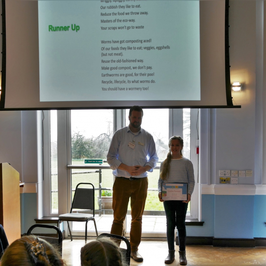 Alex Kittow stands alongside Lydia Farmer who is holding her award for her winning poem. They are both smiling at the camera. Carys' poem is displayed on a screen behind them.