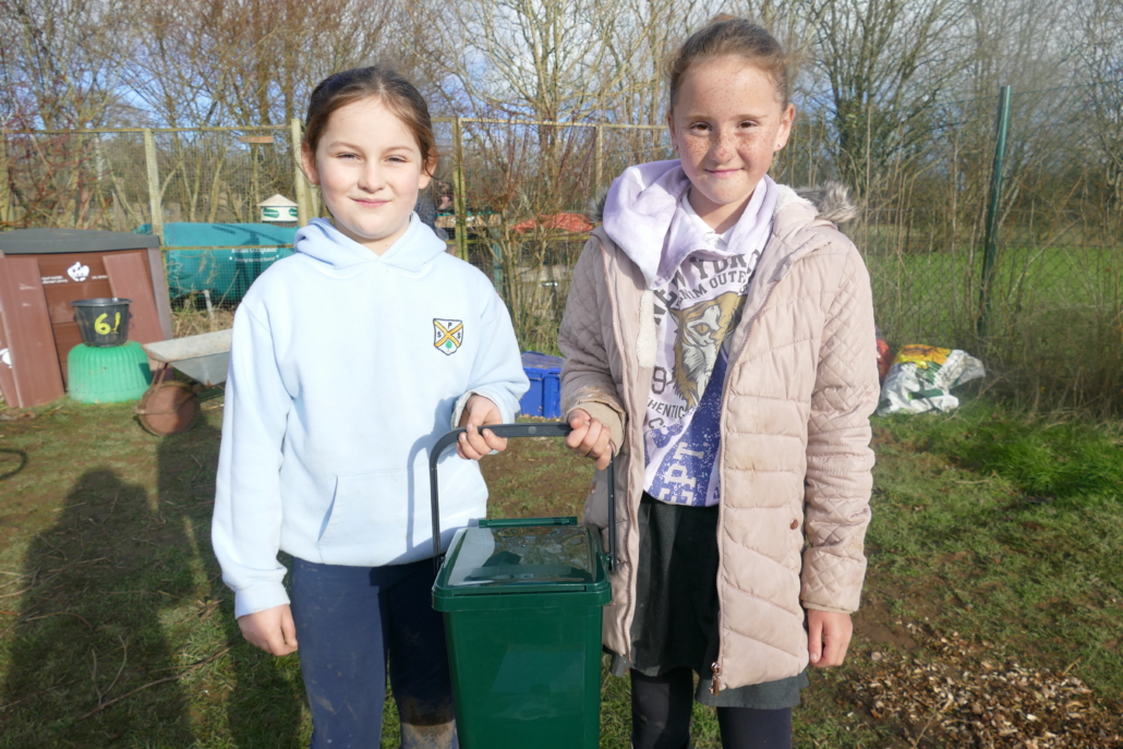 Two children holding a green compost bin standing in a school garden