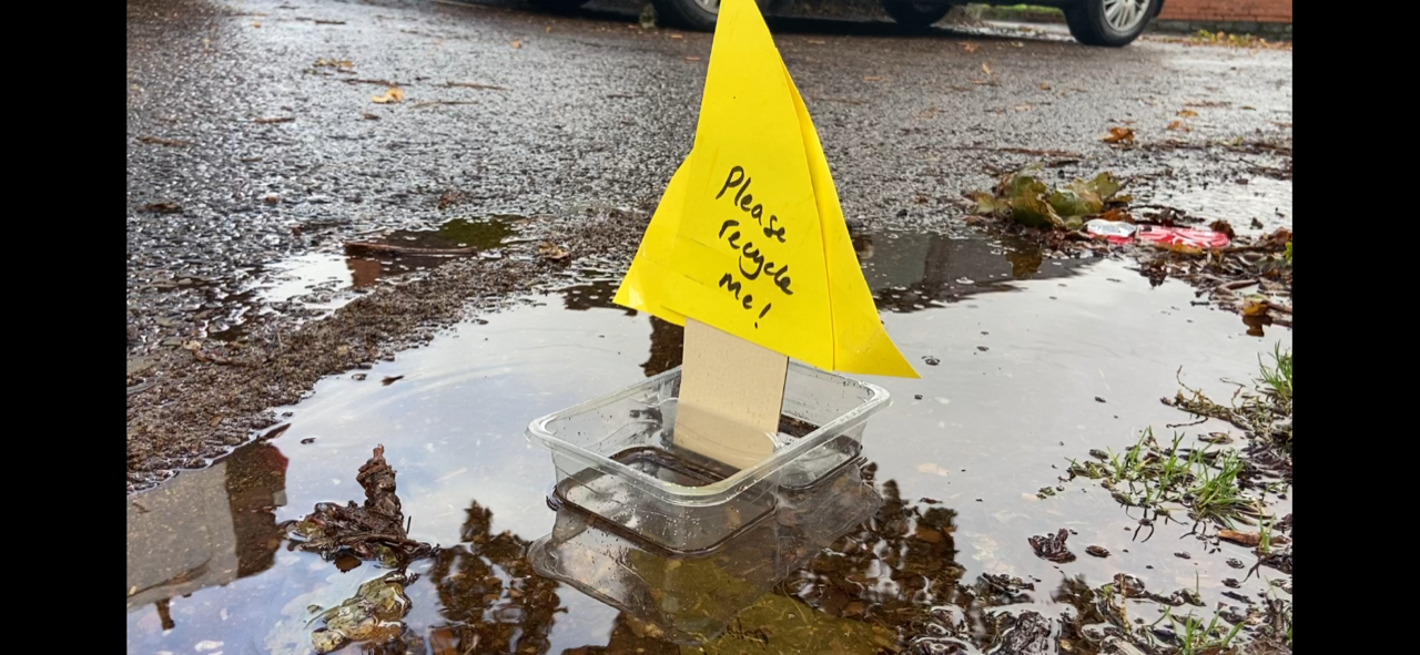 A transparent plastic pot with a piece of cardboard and papaer. The yellow paper saying 'please recycle me'. It is an example submission to the Big Recycling Hunt.