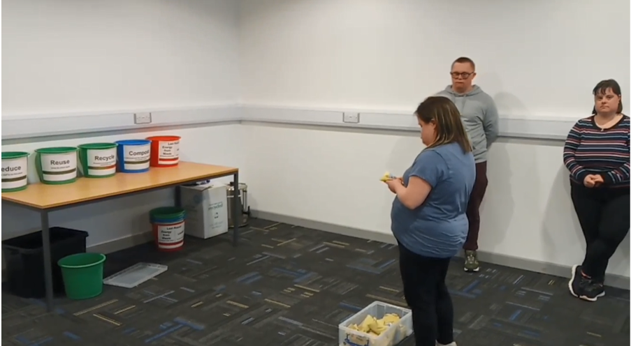 Three visitors from ROC creative, two stood against the wall and watching another visitor hold a beanbag, looking at five coloured buckets placed on a table.