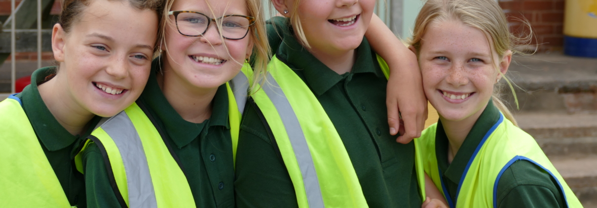 Four young girls in high-vis vests smiling at the camera, while hugging each other.
