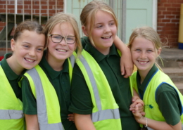 Four young girls in high-vis vests smiling at the camera, while hugging each other.