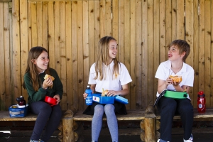 3 smiling children sitting apart in an outdoor covered seat seat eating packed lunches