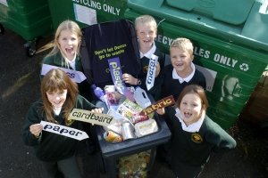 5 children around a bin filled with household rubbish holding signs that have different materials written on them.