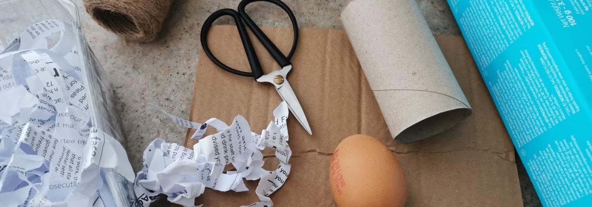 An egg is surrounded by empty cardboard packaging, a yogurt pot, a fruit punnet, shredded paper, scissors and string.