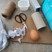 An egg is surrounded by empty cardboard packaging, a yogurt pot, a fruit punnet, shredded paper, scissors and string.