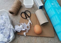 An egg is surrounded by empty cardboard packaging, a yogurt pot, a fruit punnet, shredded paper, scissors and string.