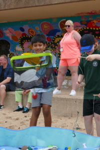 A focused young boy uses a fishing net to scoop out cardboard fishes. another smiley boy next to him uses a rope and stick to get fake fishes. This is played on Exmouth beach sands.