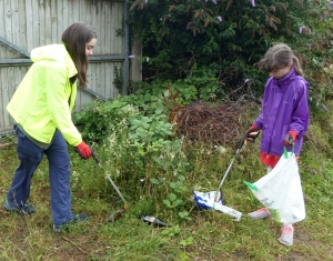 Girls wearing gloves picking up bottles and crisp packets with litter pickers from a bush.