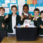 5 smiling children in school uniform standing behind a recycling box full of clean recyclable household packaging
