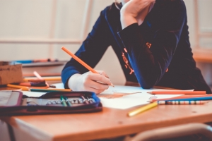 Literacy Child (no face visible, shoulders downwards) sat at desk holding a pencil crayon with pencil case, pencil crayons and paper in front