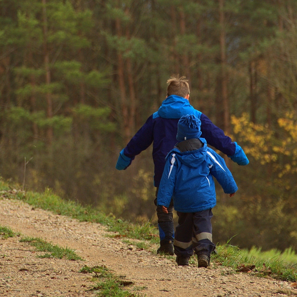 Two young children in blue coats walk on a path beside a hedge or woodland edge