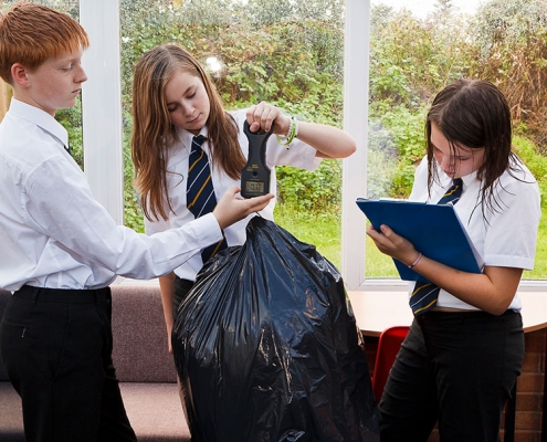 3 secondary age students weighing a rubbish bag using spring balances and clipboard