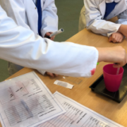 A photo of children in lab coats taking water from a cup using a pipette. This is with a recording sheet. You can't see the children's faces as its focused on their hands.