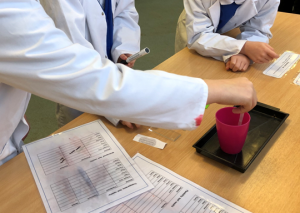 A photo of children in lab coats taking water from a cup using a pipette. This is with a recording sheet. You can't see the children's faces as its focused on their hands.