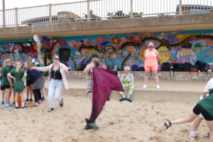 Sam is waving a scarf as he pretends to be storm, on Exmouth beach. This is in front of teachers and children who are all playing the fishing game.